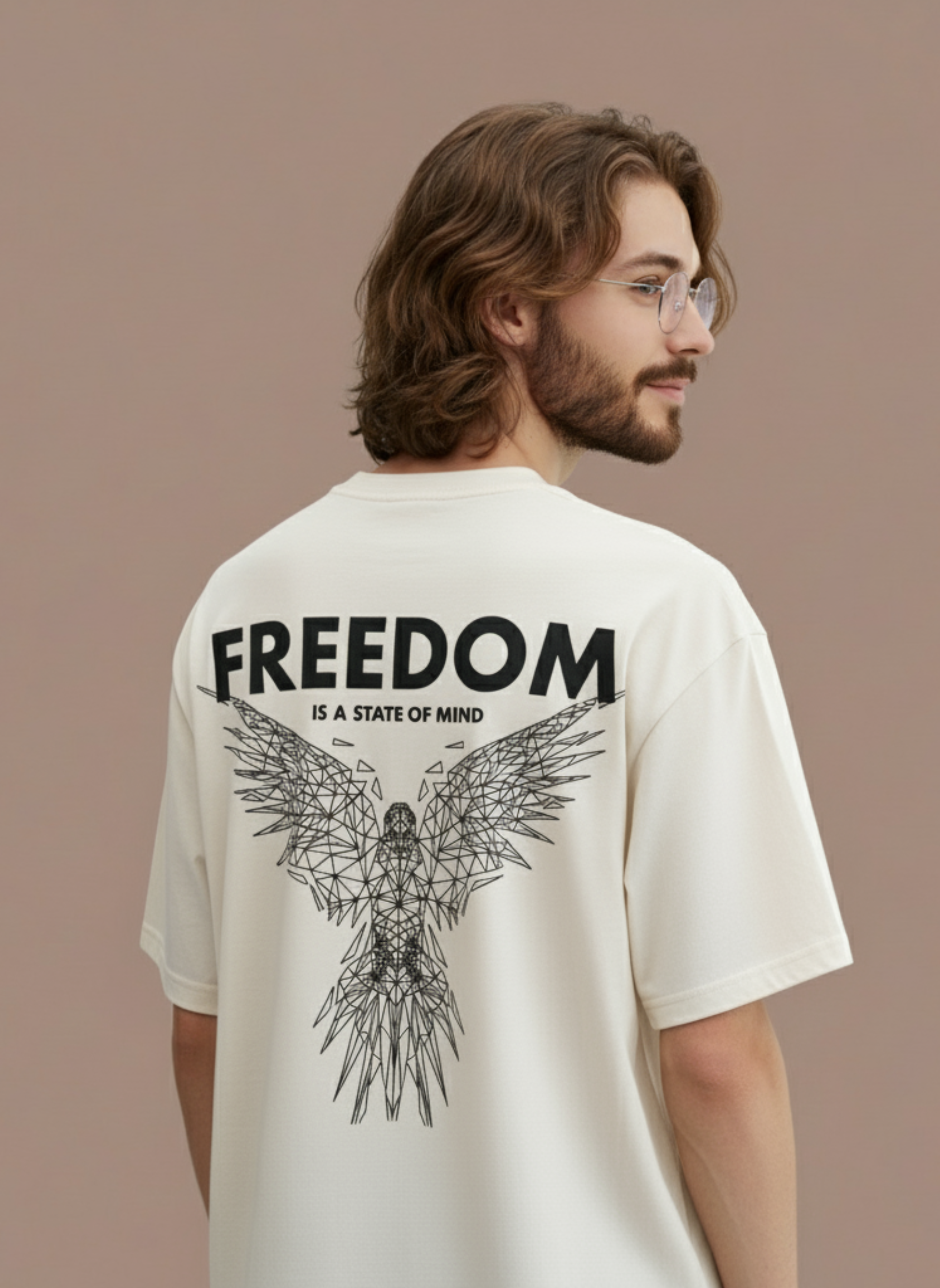 A man with wavy hair and glasses wears the VYRE Freedom Oversized Tee, a cream unisex streetwear shirt with a geometric eagle and "FREEDOM IS A STATE OF MIND" on the back, standing against a muted plain background.